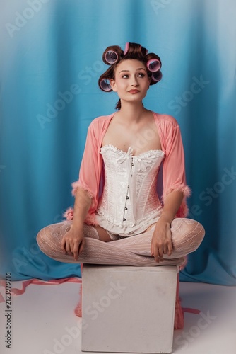 Sitting elegantly in a pastel hue, a model embraces vintage glamor with hair rollers and a flowing robe