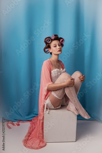 Sitting elegantly in a pastel hue, a model embraces vintage glamor with hair rollers and a flowing robe