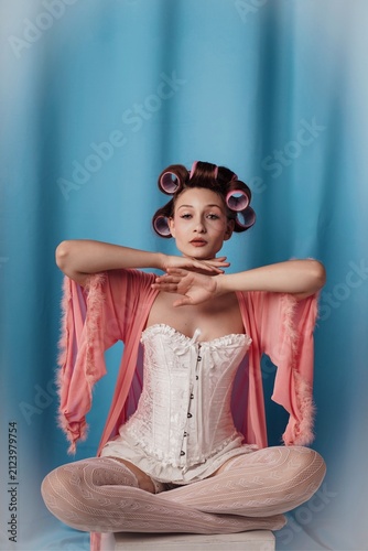 Sitting elegantly in a pastel hue, a model embraces vintage glamor with hair rollers and a flowing robe