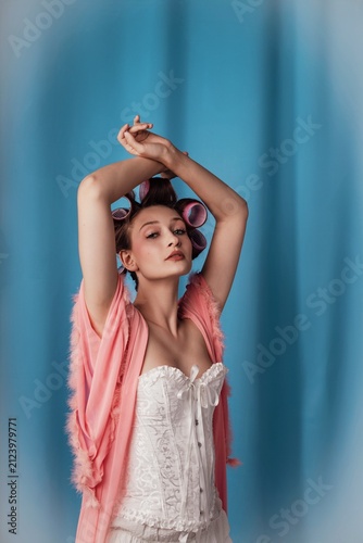 Woman posing with arms raised, wearing pink robe and hair rollers in a studio with blue background