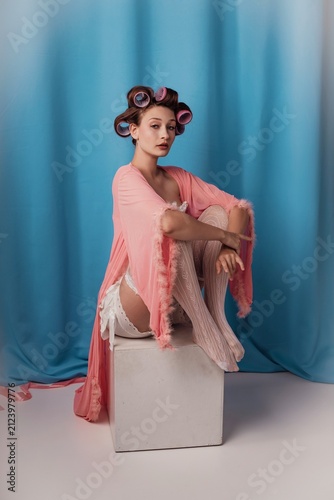 Sitting elegantly in a pastel hue, a model embraces vintage glamor with hair rollers and a flowing robe