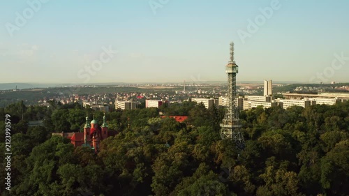 Petrin Lookout Tower rises above green trees beside Saint Lawrence Church in Prague. Historic landmark stands on hill above city background. European travel destination offers rich culture.