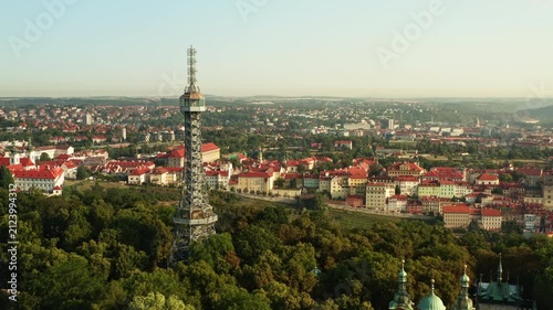 Petrin Lookout Tower stands above green forest on Petrin Hill in Prague. Historic city panorama with red tile roofs stretches under sunny sky. European capital landmark for travel concept.