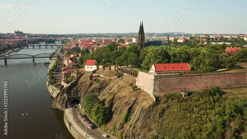 Vysehrad fortress and Basilica of St. Peter and Paul stand on a cliff in Prague. A tram enters a road tunnel below historic brick walls along the Vltava riverfront in European capital city.