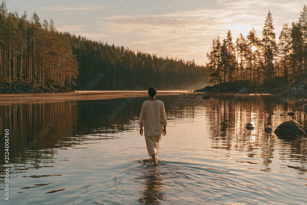 Obraz premium Peaceful figure standing in a calm lake at sunset with forest beyond, creating a serene landscape of reflection, solitude and connection with nature.