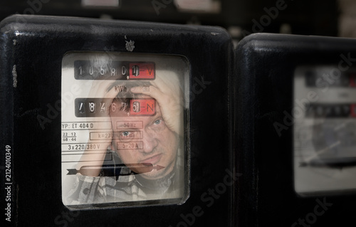 Electrical meter with reflection of shocked customer reading electricity consumption. Close-up stressed man with head in hands checking electric power measuring device at high energy prices. Business.