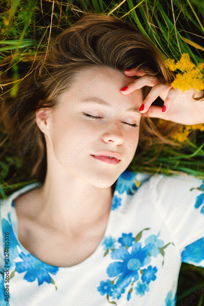 Naklejka premium Beautiful smiling cute blonde young woman lies in green grass with eyes closed, red nails resting on her forehead, wearing a floral dress and enjoying a peaceful summer moment outdoors