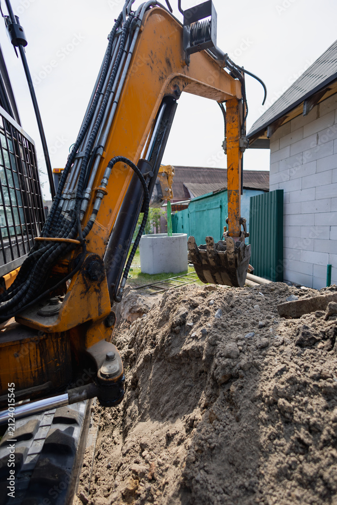 Fototapeta premium A digger works in a backyard, creating a trench for construction. Soft earth is piled high, and nearby structures are visible under a clear sky
