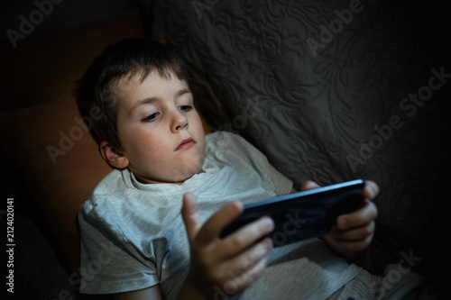 A 6-7-year-old boy plays on his mobile phone and checks social media while lying down instead of sleeping. Digital addiction in children, including decreased concentration and harm to mental health.