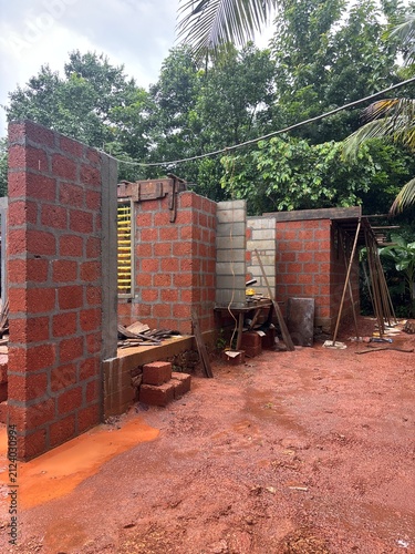 A residential house construction site in Kerala, India, featuring reinforced concrete columns, steel reinforcement bars (rebar), foundation work, and partially completed masonry. The image showcases m