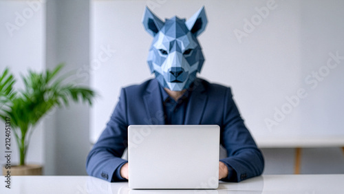 Blurred man in a suit wearing a low-poly blue wolf mask, sitting at a white desk in a minimalist office with a potted plant, typing on a laptop with a softly background. Focus foreground laptop.