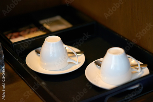 Two white ceramic coffee cups placed upside down on matching saucers inside a black service tray.