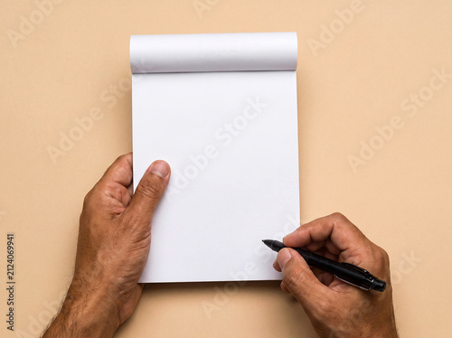 Person holding a vertical notepad and writing with a pen on a beige background