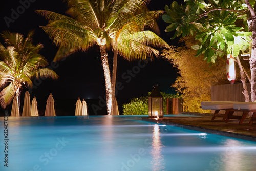 Beautiful illuminated resort swimming pool at night surrounded by tall palm trees and warm glowing lights under the dark sky.