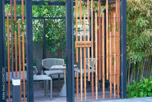 Modern outdoor seating area with comfortable chairs, a glass table, and stylish vertical wooden slats surrounded by green plants for smoking area