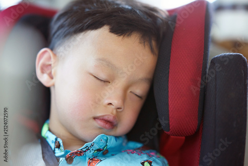 A close-up shot of a young Asian boy sleeping peacefully while buckled safely inside a red and black child safety car seat.