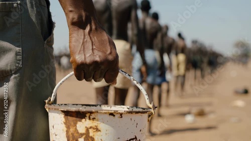 Hand holding white rusted bucket with people standing in line outdoors, close up shot