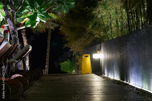 Beautifully illuminated wooden walkway path at night surrounded by lush tropical green plants and trees.