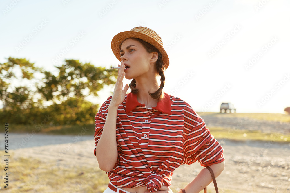 Obraz premium Young woman in a straw hat and striped shirt yawns while standing outdoors. Authentic lifestyle portrait of a girl posing with braided hair on a sunny summer day.