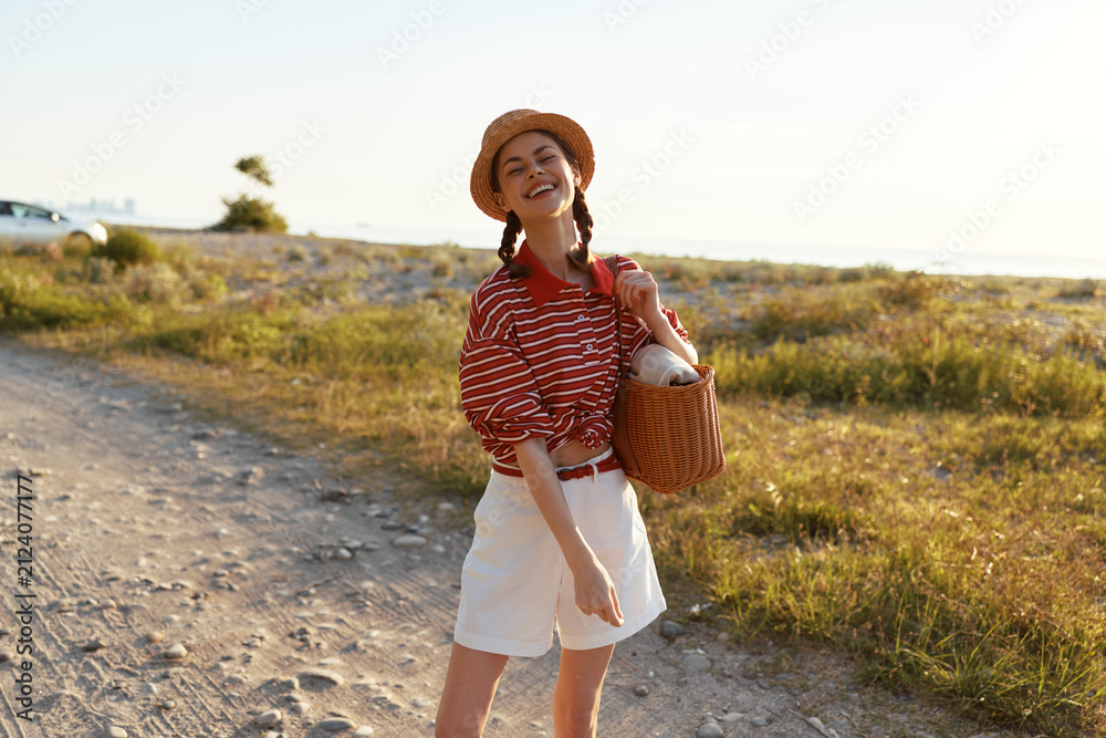 Obraz premium Happy woman enjoying lifestyle during a summer walk on a sandy beach. She wears a straw hat and striped shirt, holding a bag. Sunny outdoor scene with warm lighting.