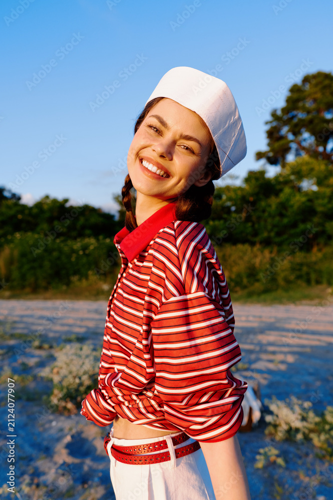 Obraz premium Smiling woman outdoor lifestyle portrait in a striped shirt and beret. The joyful girl poses on a beach at sunset. Concept of summer leisure, happiness, and authentic style.