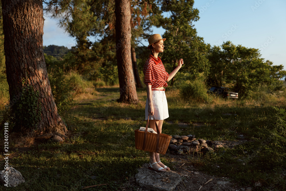 Obraz premium Young woman standing in forest holding wicker basket during sunset. Lifestyle portrait in nature with warm golden light. Concept of summer picnic recreation outdoor.