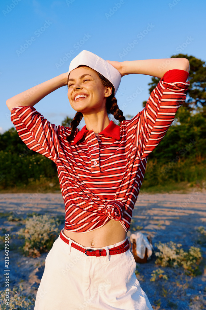 Obraz premium Smiling woman lifestyle posing outdoors in a striped shirt and hat. Sunny summer background, golden hour lighting. Concept of travel, adventure, freedom, and vacation mood.