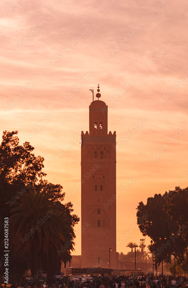 Obraz premium Tall minaret tower rises above crowded market below in the ornage light of sunset, Marrakesh Morocco
