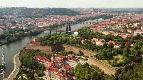 Historic Vysehrad fortress overlooks Vltava river and city bridges in Prague. European architectural landmark serves as popular tourist attraction. Scenic urban landscape represents travel.
