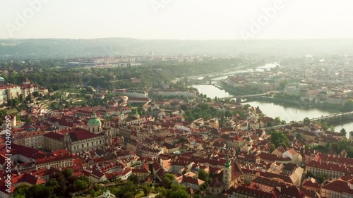 Early morning sun illuminates Prague cityscape and Vltava river bridges. Historic European architecture showcases red tiled roofs. Soft morning light creates peaceful travel destination.
