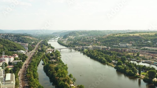 Vltava river flows through green urban landscape in Prague. Scenic riverside displays road traffic and distant bridges. Natural city environment serves as European tourism destination.