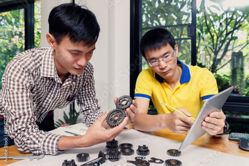 Two Asian engineers discussing mechanical design with metal gears and digital tablet in office. Teamwork and industrial machine development concept.