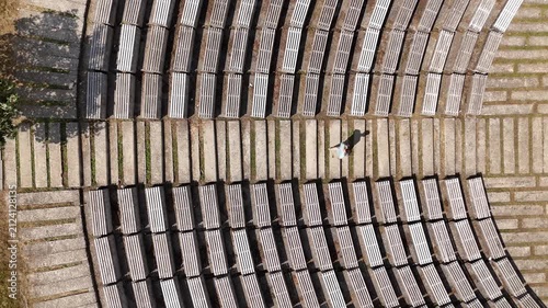 Top down aerial view of a lonely woman walking through an empty outdoor amphitheater, with symmetrical wooden benches and concrete steps casting long shadows on a sunny day