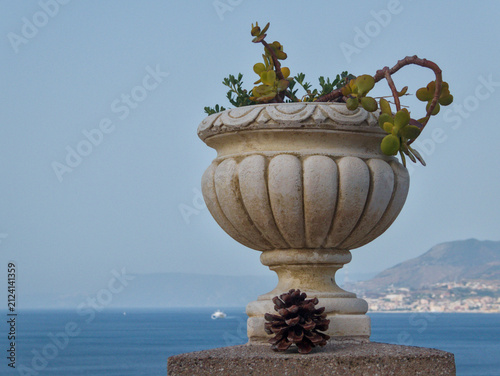 Canvastavla Classic stone urn planter with succulent plant and a pinecone resting on a stone balustrade over scenic blue Mediterranean sea view in Messina, Sicily, Italy during sunny summer day