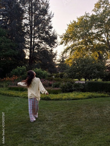 A woman walks through a peaceful green park surrounded by towering trees, embracing calm, mindfulness, and the refreshing beauty of nature during a relaxing outdoor escape.