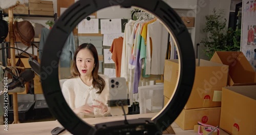 A stylish Asian woman fashion designer, influencer livestreams from studio workspace, Promotes her brand and communicates with online customers on social media platform. entrepreneurship, e-commerce.