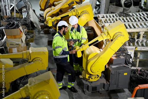 Workers inspect robotic arms in a factory during day hours while discussing technology and safety protocols