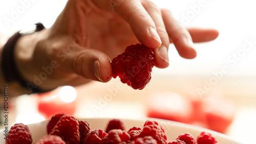 Human hand holding a fresh red raspberry on an open palm over a white bowl filled with ripe berries.