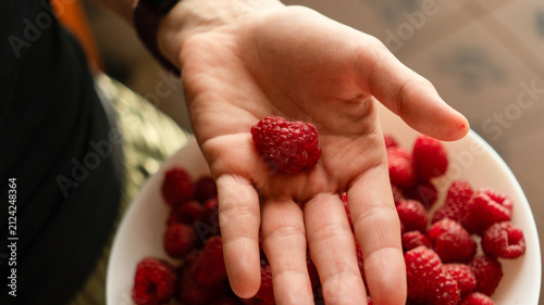 Human hand holding a fresh red raspberry on an open palm over a white bowl filled with ripe berries.