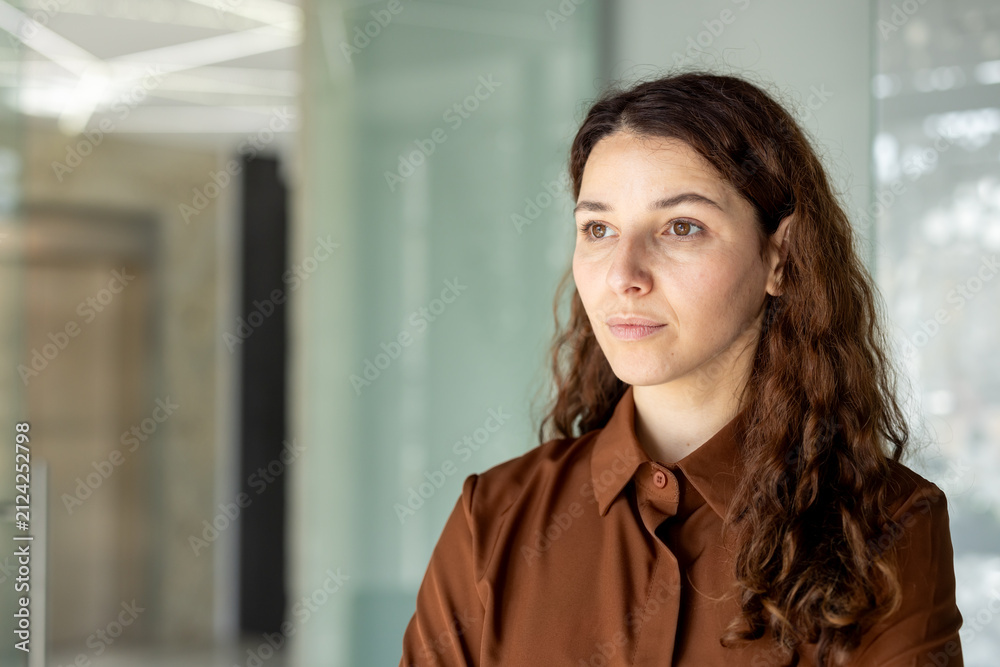 Obraz premium Woman with long brown hair wearing a shirt, looking away thoughtfully while standing in a bright corporate office interior, representing focus, success, and forward-thinking