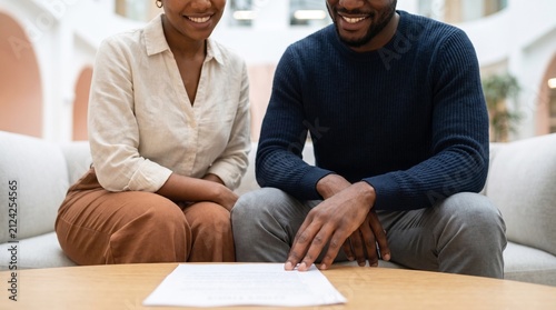 Happy Black couple reviewing mortgage contract or financial loan agreement at home, smiling young family signing real estate agreement document for modern house purchase concept.