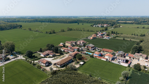 Rural Hamlet Among Fields: An aerial view reveals a picturesque rural village nestled amidst vibrant green farmland under a clear blue sky, showcasing a tranquil countryside scene.