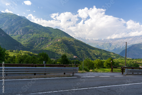 Fotografía A scenic mountain highway with guardrails overlooks a lush green valley and dramatic forested peaks in Italy's Dolomites