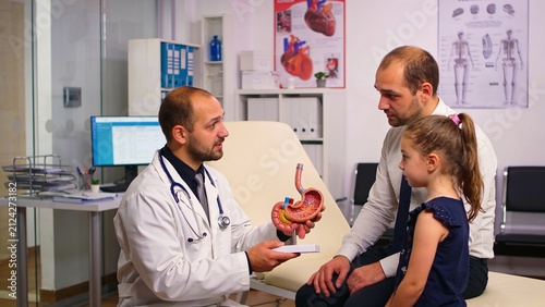 Gastroenterologist explaining stomach model to father and daughter during consultation in modern medical clinic.