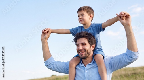 Father carries son on shoulders outdoors, both smiling against a clear blue sky with grassy field in the background