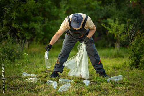 A person is bending down to pick up plastic bottles from the ground. They are using a bag to collect the waste. The sunny day shows a clean, green area with trees in the background