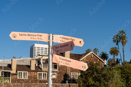 Tourist wayfinding sign in Santa Monica, California showing directions to iconic coastal attractions. Blue sky, palm trees and city buildings create a classic Southern California travel atmosphere