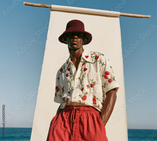 Stylish male fashion model posing on a beach studio backdrop in a floral shirt, bucket hat and bold red shorts, blending coastal lifestyle with contemporary editorial summer fashion.