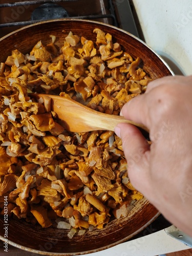 Fresh chanterelle mushrooms frying in a pan while a female hand stirs with a wooden spatula, homemade rustic cooking