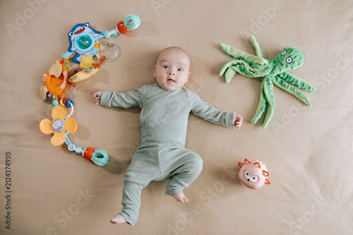 Cute 3 month baby boy lying among various playful colorful toys on beige blanket. Infant surrounded by vivid objects participating actively in sensory exploration and learning. 